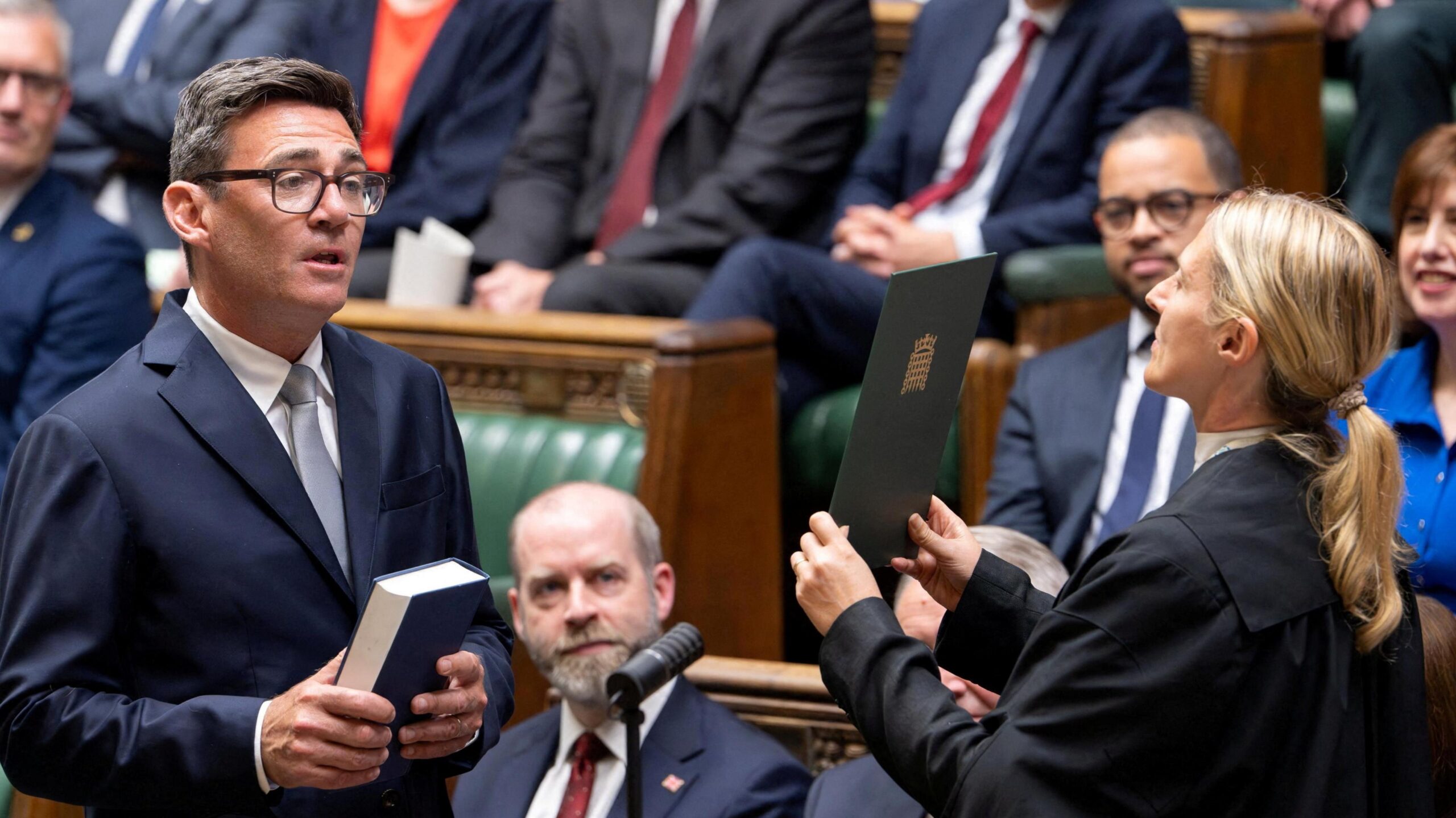 Andy Burnham, in a suit and tie, holds a Bible and reads from a card held up by a female Commons official in the House of Commons chamber