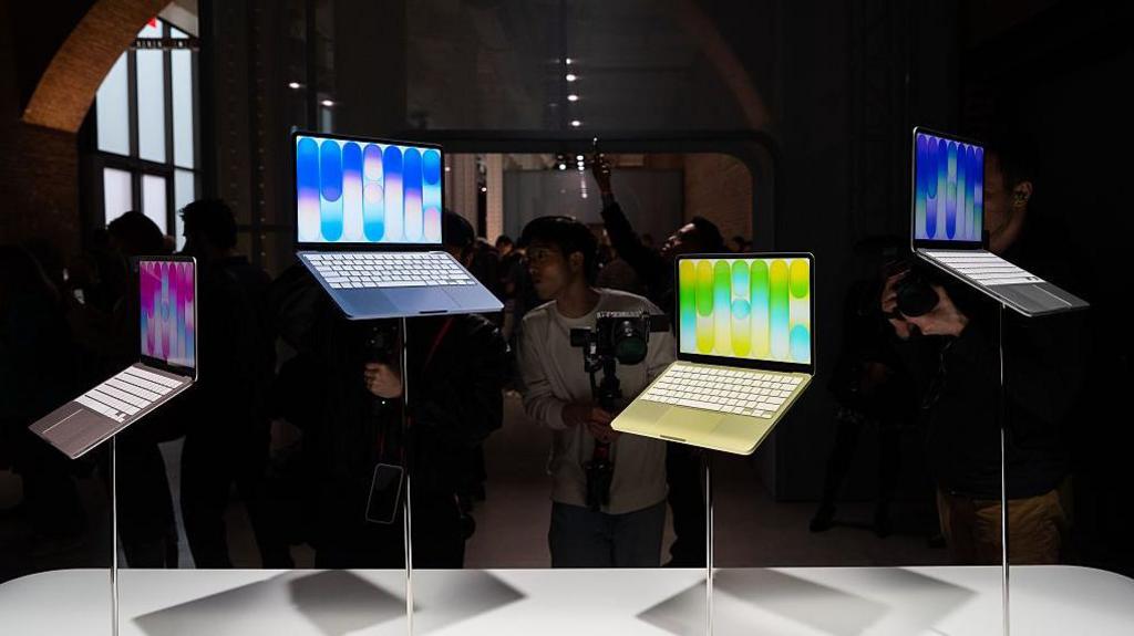 A set of colourful Apple Neo laptops positioned at varying heights on stands in a display at an Apple Store, with photographers crowding around to capture them images of them.