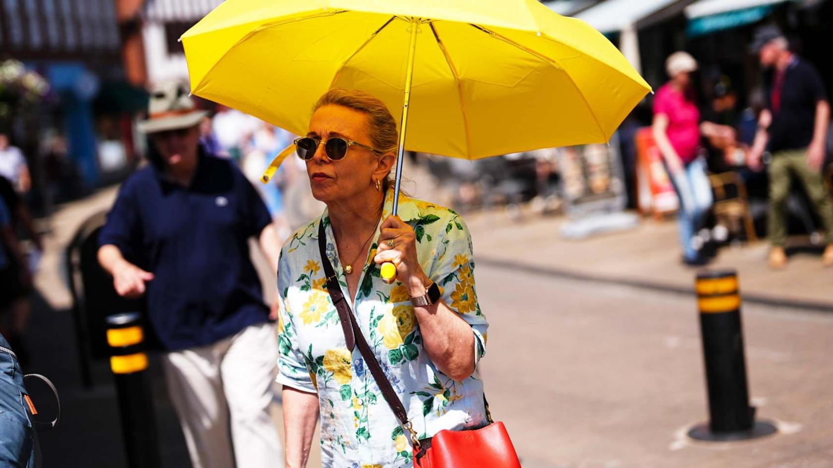 A women holds a yellow umbrella to shade her from the sun. She walks along a high street in the UK wearing a summer dress and carrying a red handbag.  A man behind her is wearing a hat and a dark top.