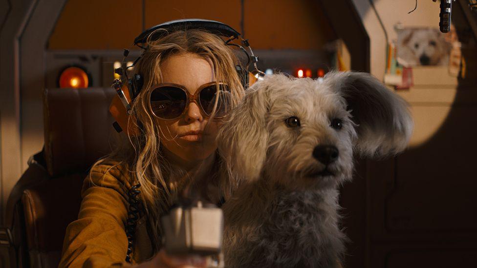 A scene from Supergirl shows the title character sitting in a chair, wearing large, vintage over-ear headphones and large sunglasses in a darkened room. Her adorable white terrier sits on her lap looking alert.