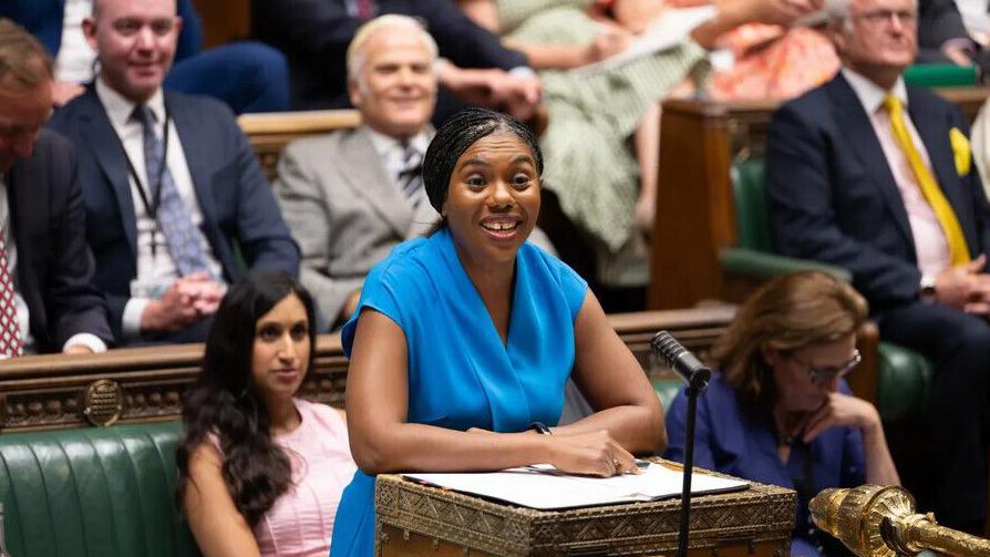 Kemi Badenoch in the House of Commons at the despatch box. She is wearing a blue dress and behind her are Conservative MPs.