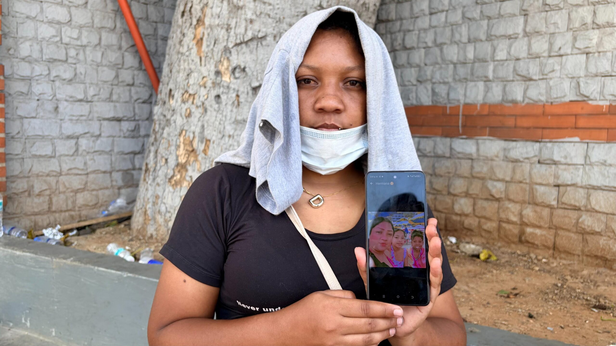 A woman with a grey t-shirt covering her head holds up a mobile phone to display a photo of her with her two teenage daughters.