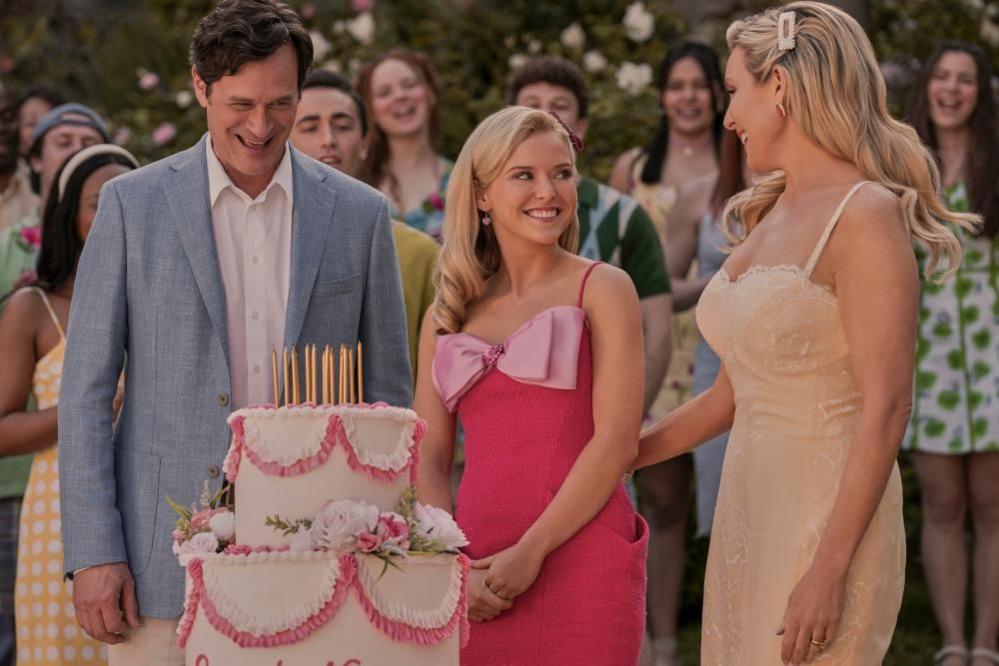 A man and two women smiling at each other, standing infront of a two-tierd cake, with gold candles and pink decorations. People are in the background, also smiling.