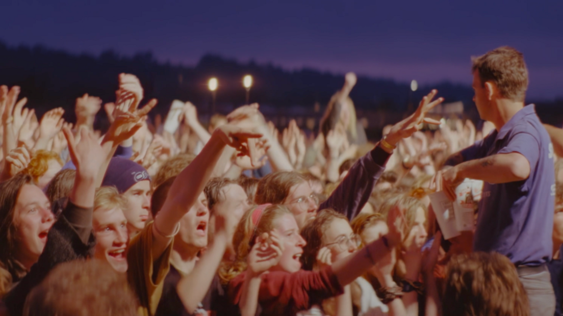 A close up of a group of festivalgoers at night, lit up from the stage, all with their hands in the air, and some are reaching towards drinks, which a stage-hand is passing to them