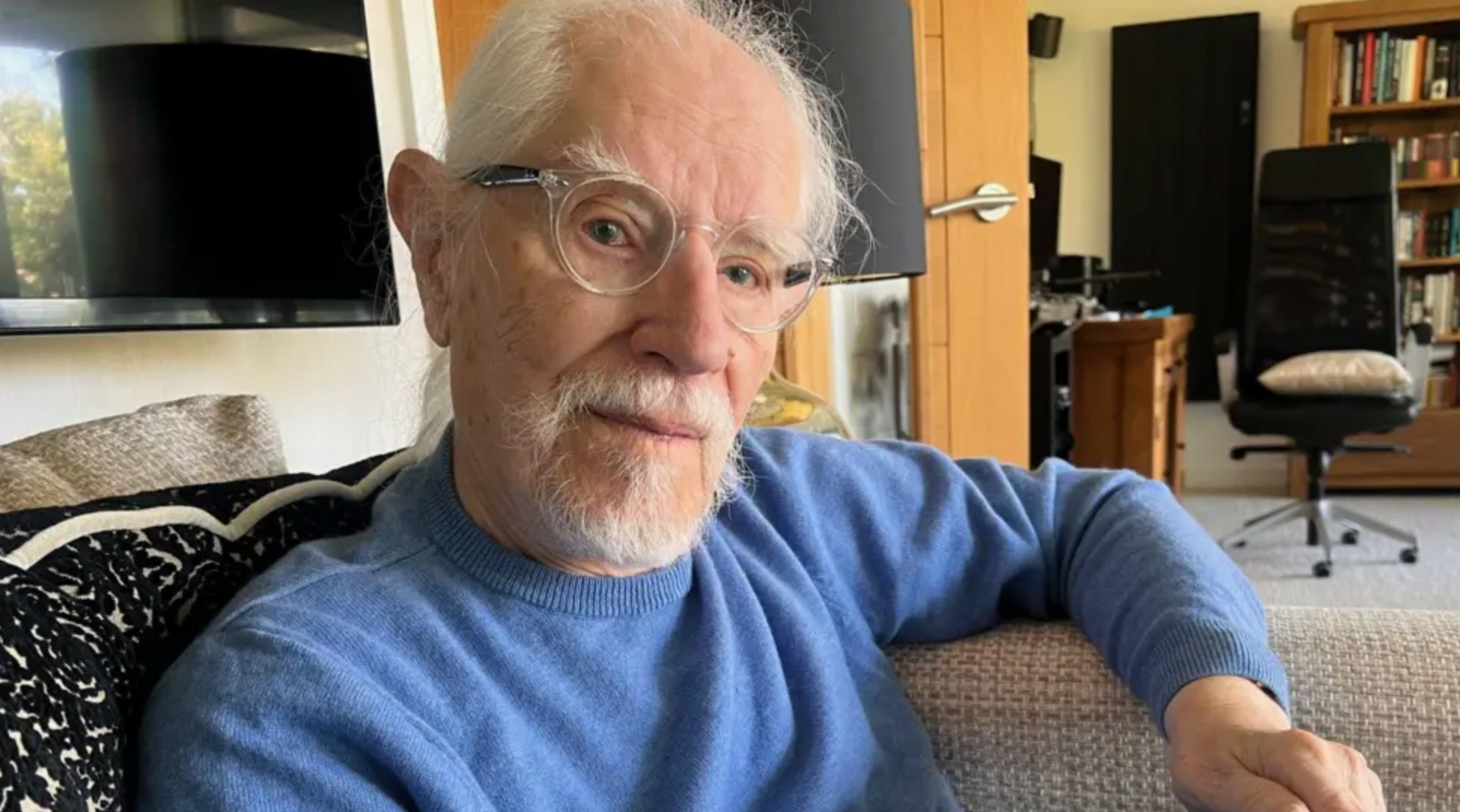 A man wearing a blue jumper and glasses looks directly into the camera as he sits on a sofa in a living room