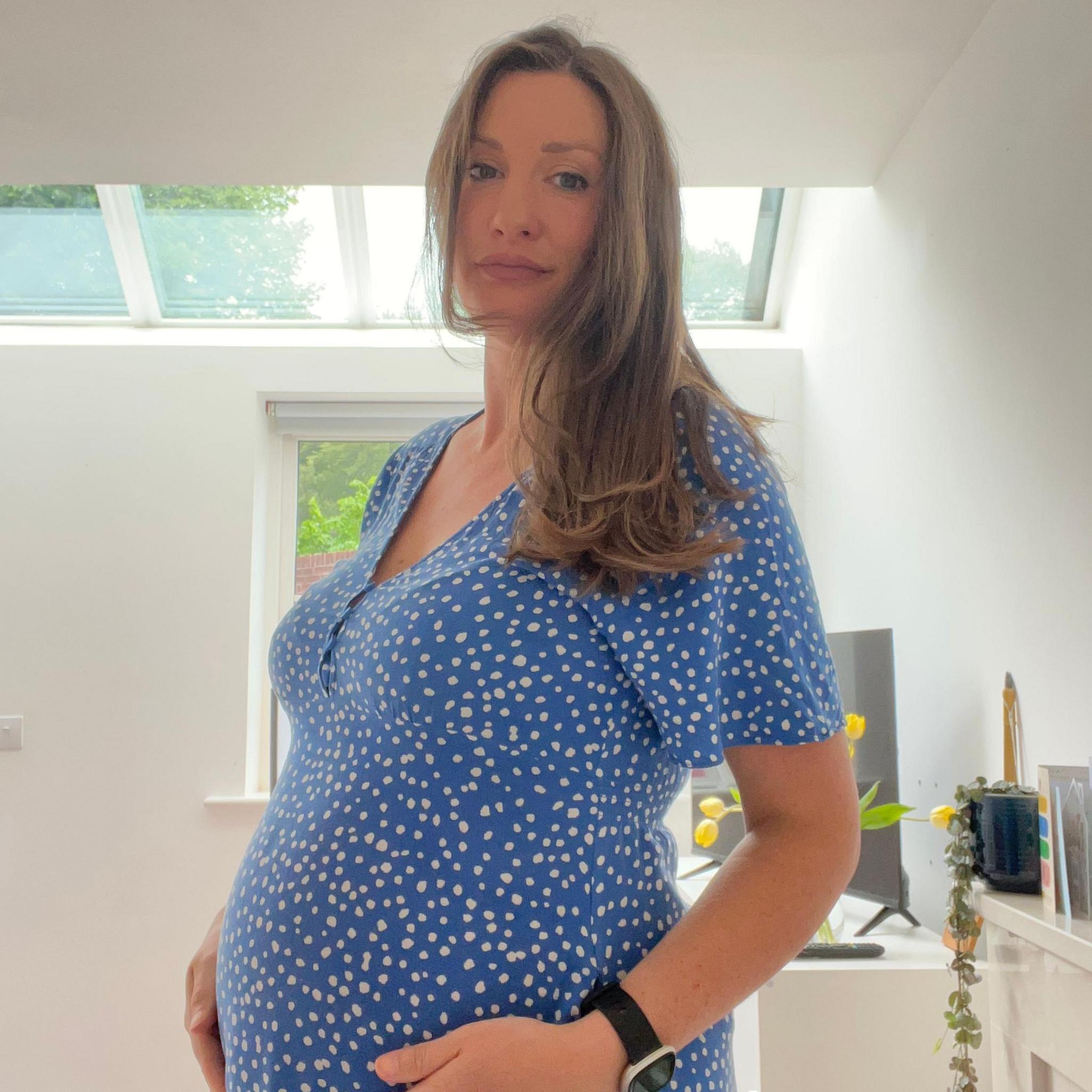 A pregnant woman standing up in a room with white walls and a window behind her. She has long light brown hair and is wearing a blue dress and white dots.