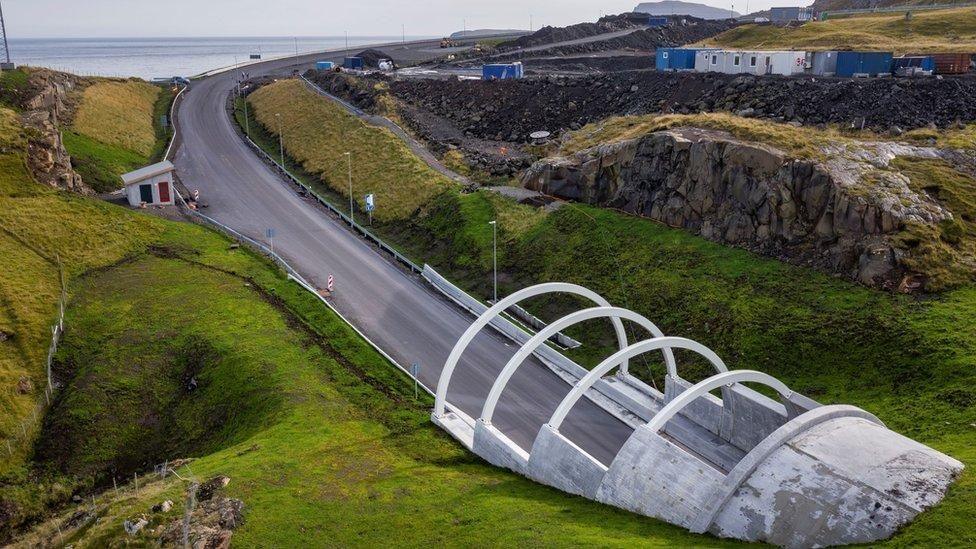 Aerial view of a modern road bridge with four white arch supports crossing a grassy ravine near a rocky coastline. The newly paved road curves toward the sea, with rugged cliffs, scattered buildings, and open water visible in the background.