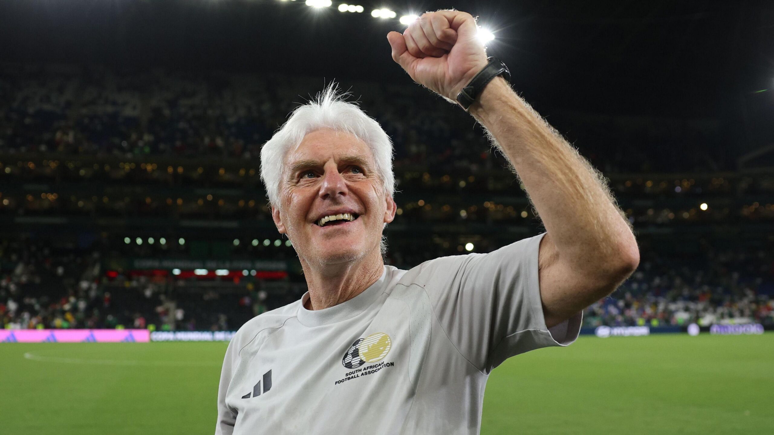 Hugo Broos, visible from chest up wearing a grey South Africa training top, smiles and holds up his left fist in celebration. He stands in front of a football pitch as full stands in a stadium can be seen out of focus in the background