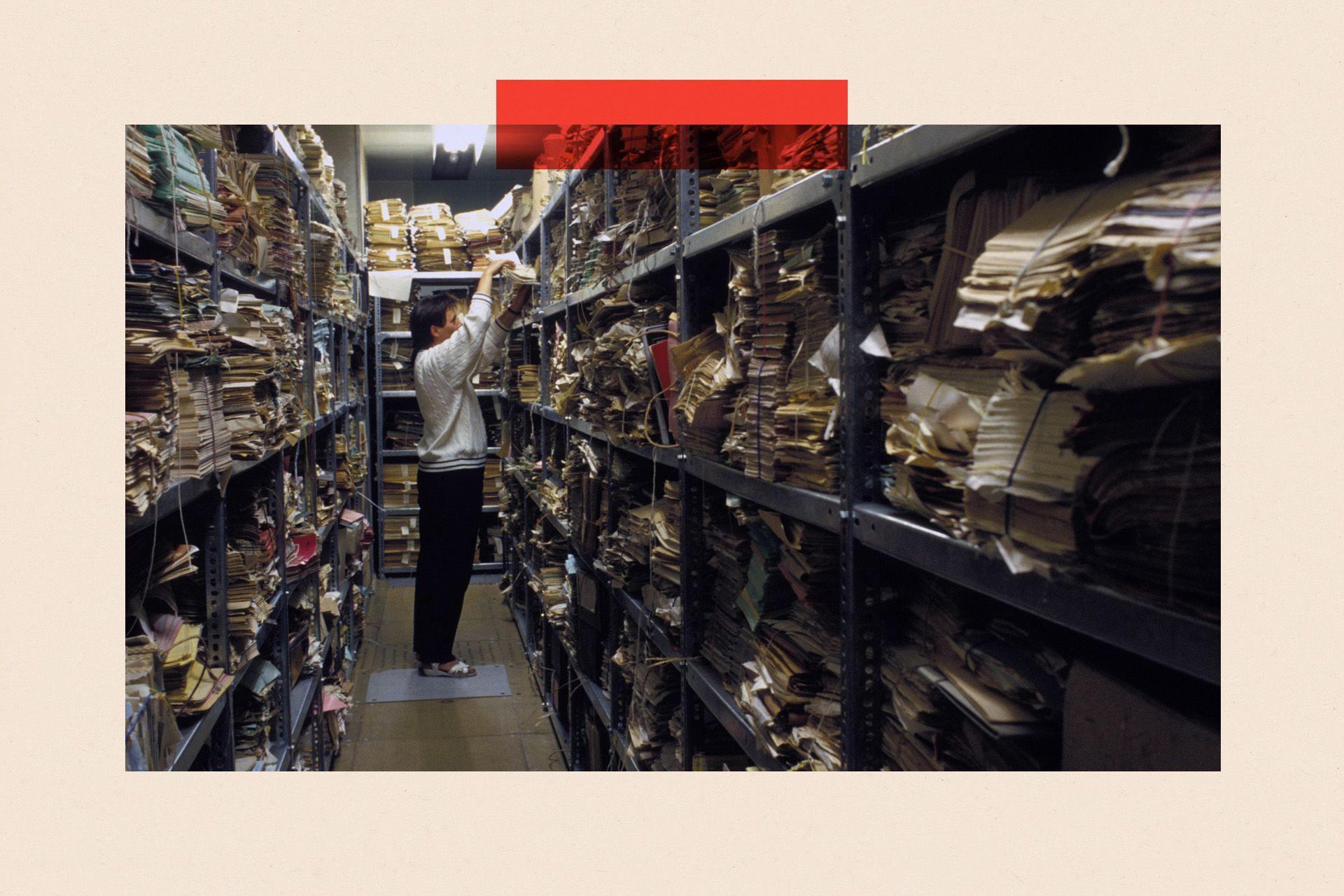 A man in a light grey sweatshirt pulls a stack of files off a shelf in an archive room, which is filled with other files