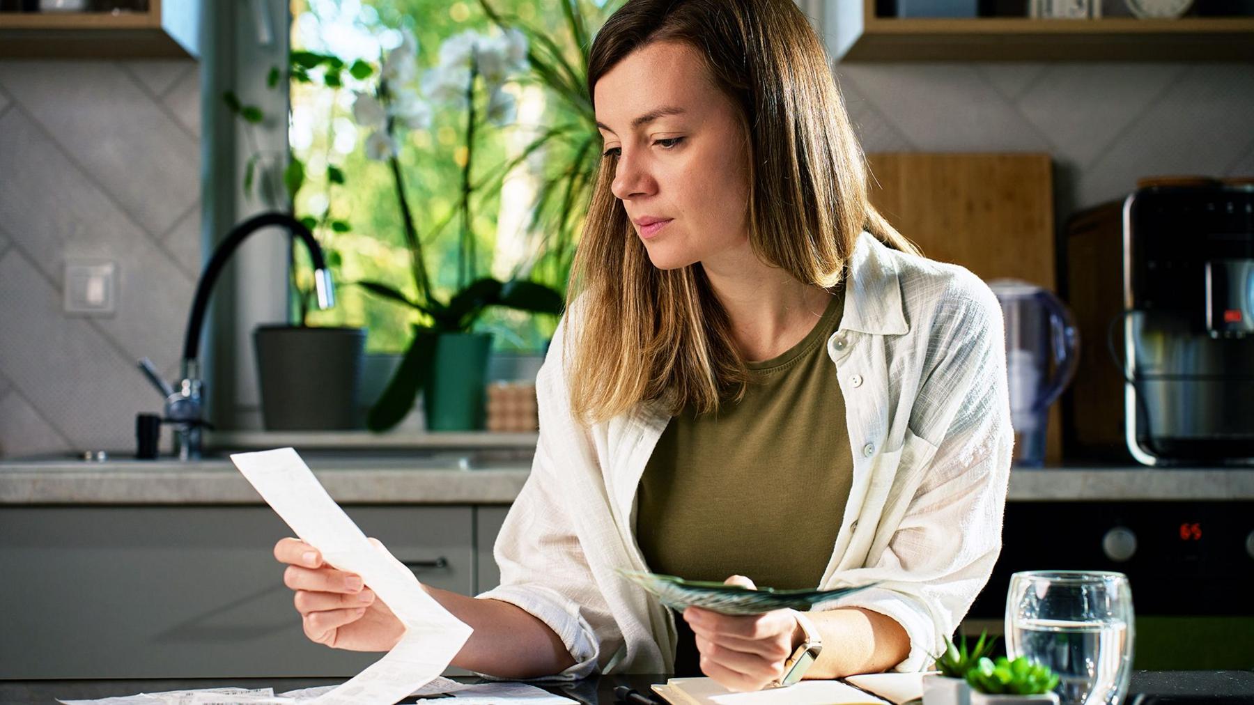 A woman sitting at a table and  looking at houshold bills in her kitchen . In the background is a window showing  green plants and trees