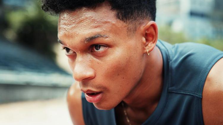 A young man, wearing a sports vest top, is out for a run. He is hunched over, hands on knees, sweating.