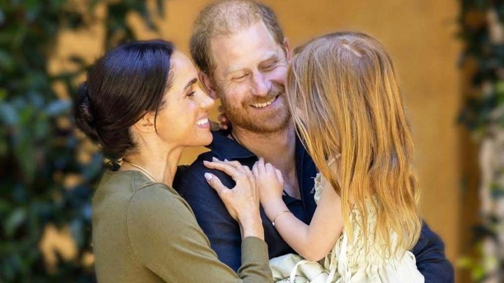 Duke and Duchess of Sussex in casual outfits embracing and holding Lilibet in a summer dress, she has bare feet and her back is to the camera; she has long, red hair