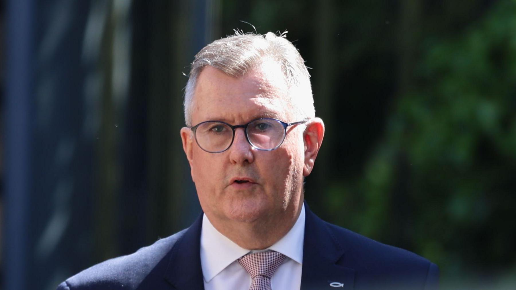 A man with short grey hair and rounded glasses is standing outside, with some out of focus trees behind him. The man is dressed in a dark suit jacket with white shirt and light pink tie, which has a textured pattern. There is a small silver fish symbol attached to the lapel of his jacket.
