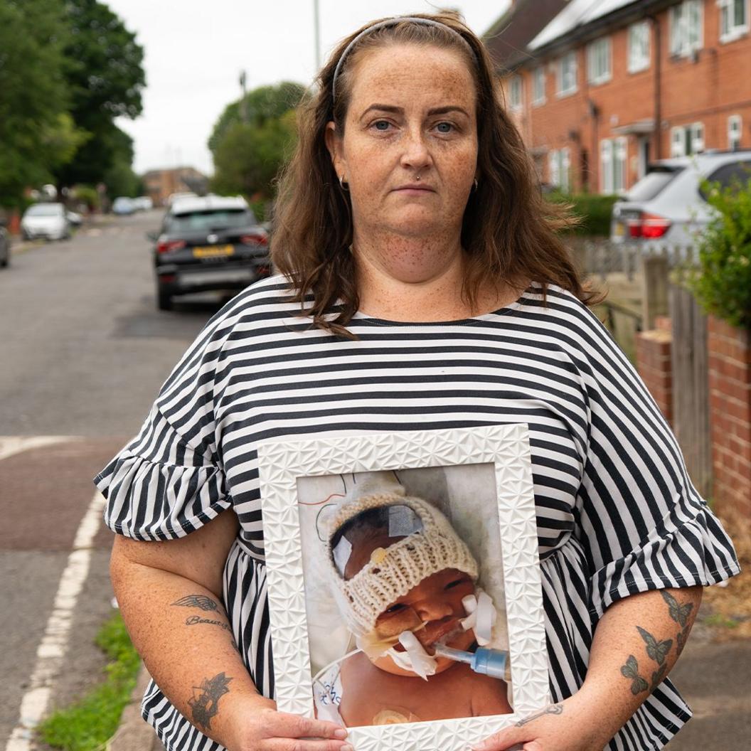 Hayley Coates, in a striped top, stands on a street holding a picture of Kaylan