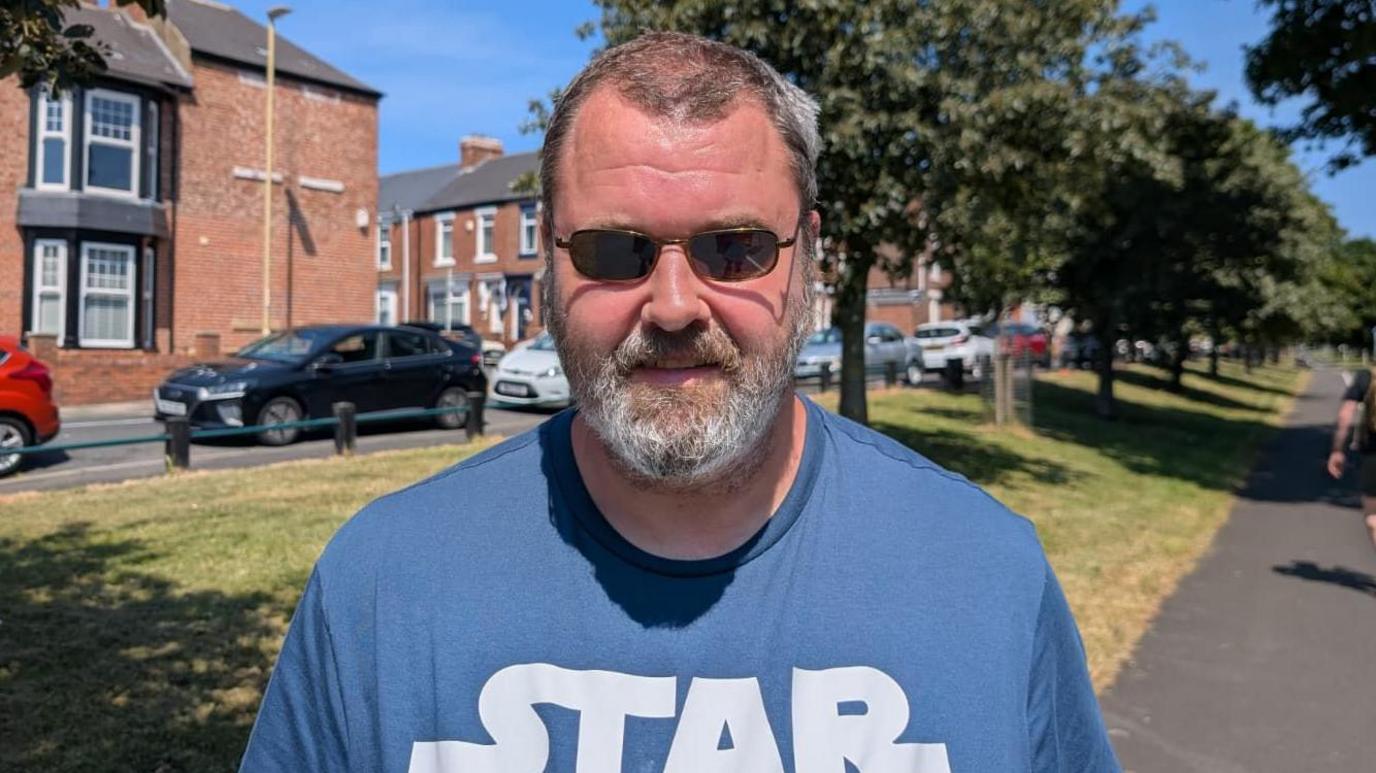 Jason who is wearing a blue Star Wars t-shirt and dark sunglasses stands in front of a leafy avenue split for walkers and cyclists. In the background are blocks of terraced houses and parked cars.