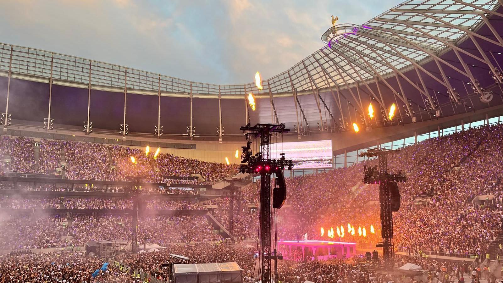 The stadium at dusk. Bad Bunny is performing on top of a small home, illuminated in pink. Fire is shooting out of the stage and lighting towers.