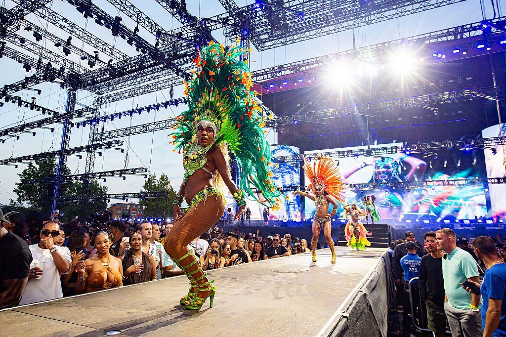 Dancers perform during day three of Wireless Festival in 2025 - they are shown wearing bright coloured green and orange costumed, on a platform with crowds either side and bright lights in the background.