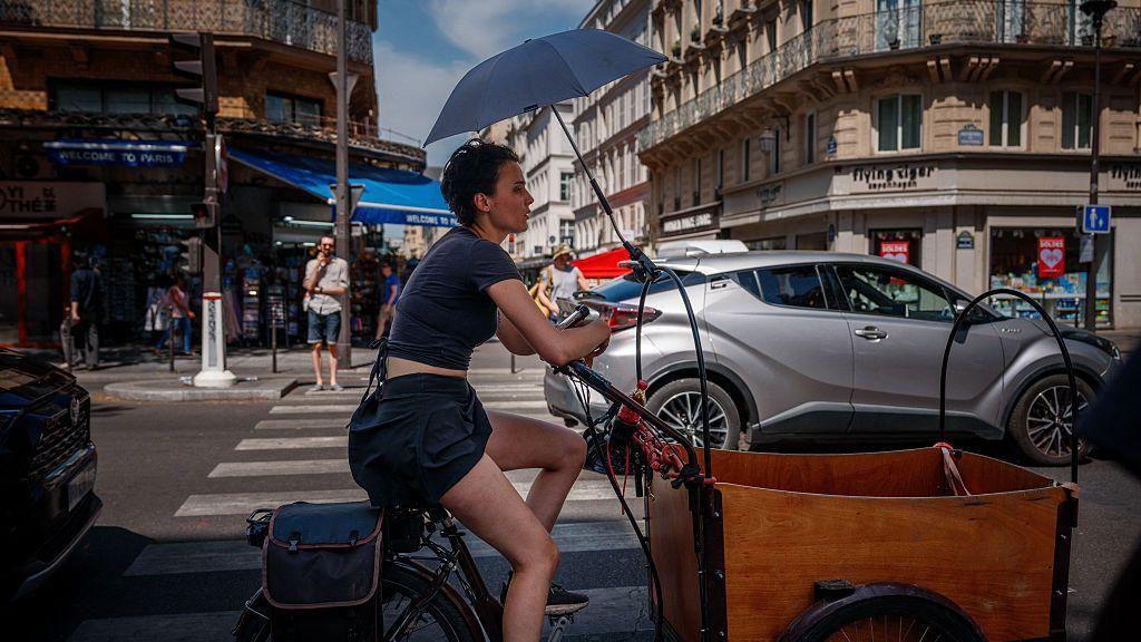 A woman protects herself from the sun under an umbrella as she rides a bicycle in Paris on June 25, 2026, as France experiences a heatwave.