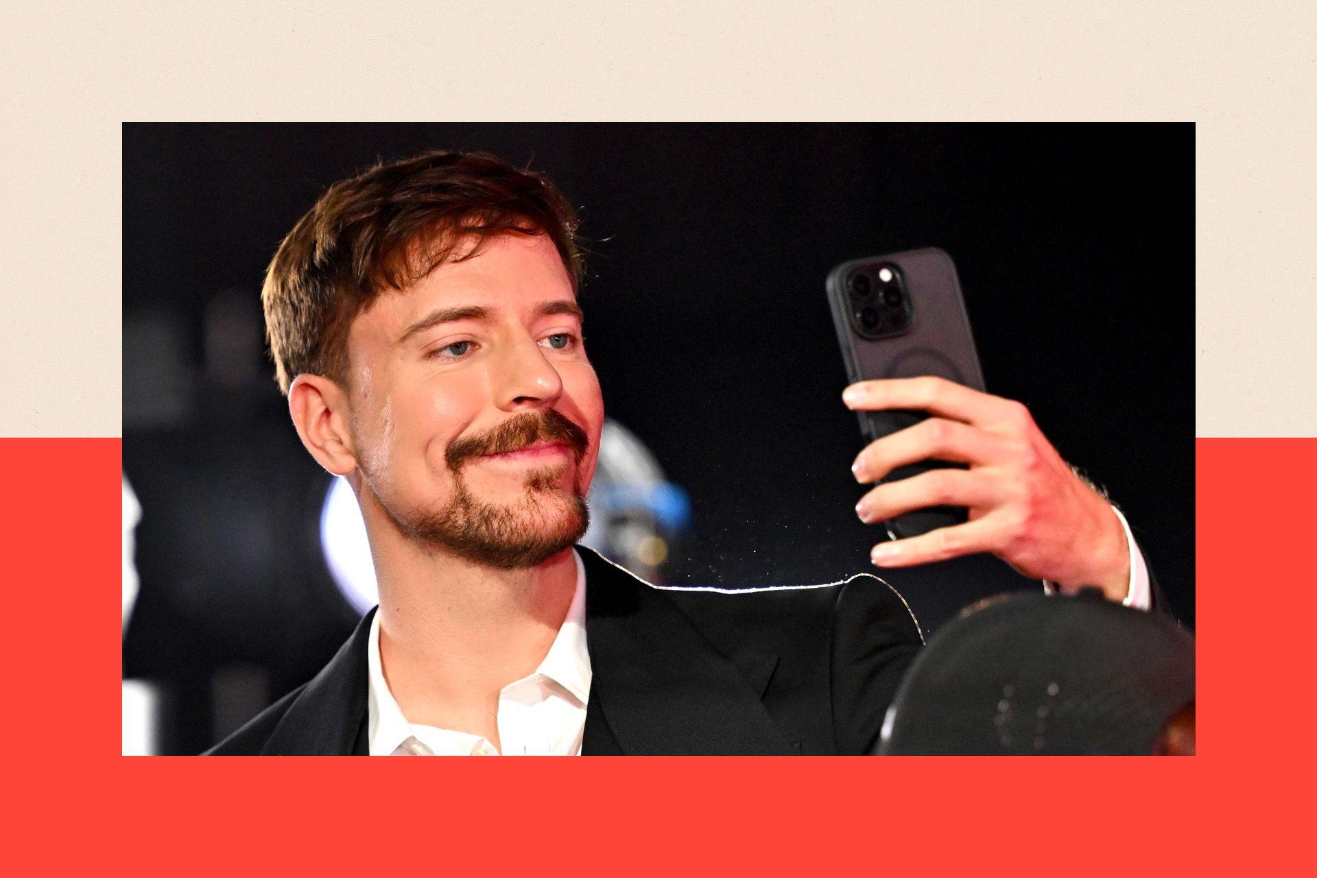 YouTube star MrBeast attends the 2025 MTV Video Music Awards. He is a white man with dark hair and a dark beard. He is wearing a dark suit jacket and a white open-necked shirt and is holiding a mobile phone in front of him as if taking a selfie.