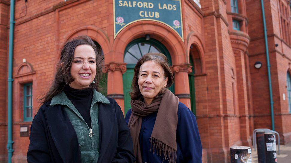 Laura Slingsby (left) chief executive at Salford Lads and Girls Club and Claudia Kenyatta of Historic England pose for a picture outside Salford Lads and Girls Club. Laura has long dark brown hair and is wearing a black polo neck under a green corduroy dress and black coat.Claudia Kenyatta has bobbed brown hair and wears a brown scarf and navy blue top with black trousers. They are both smiling.