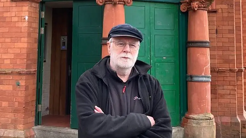 Leslie Holmes, culture and heritage projects manager at the Salford Lads and Girls Club stands outside the club. He is wearing a black cap and black top and fleece and has grey hair and beard.