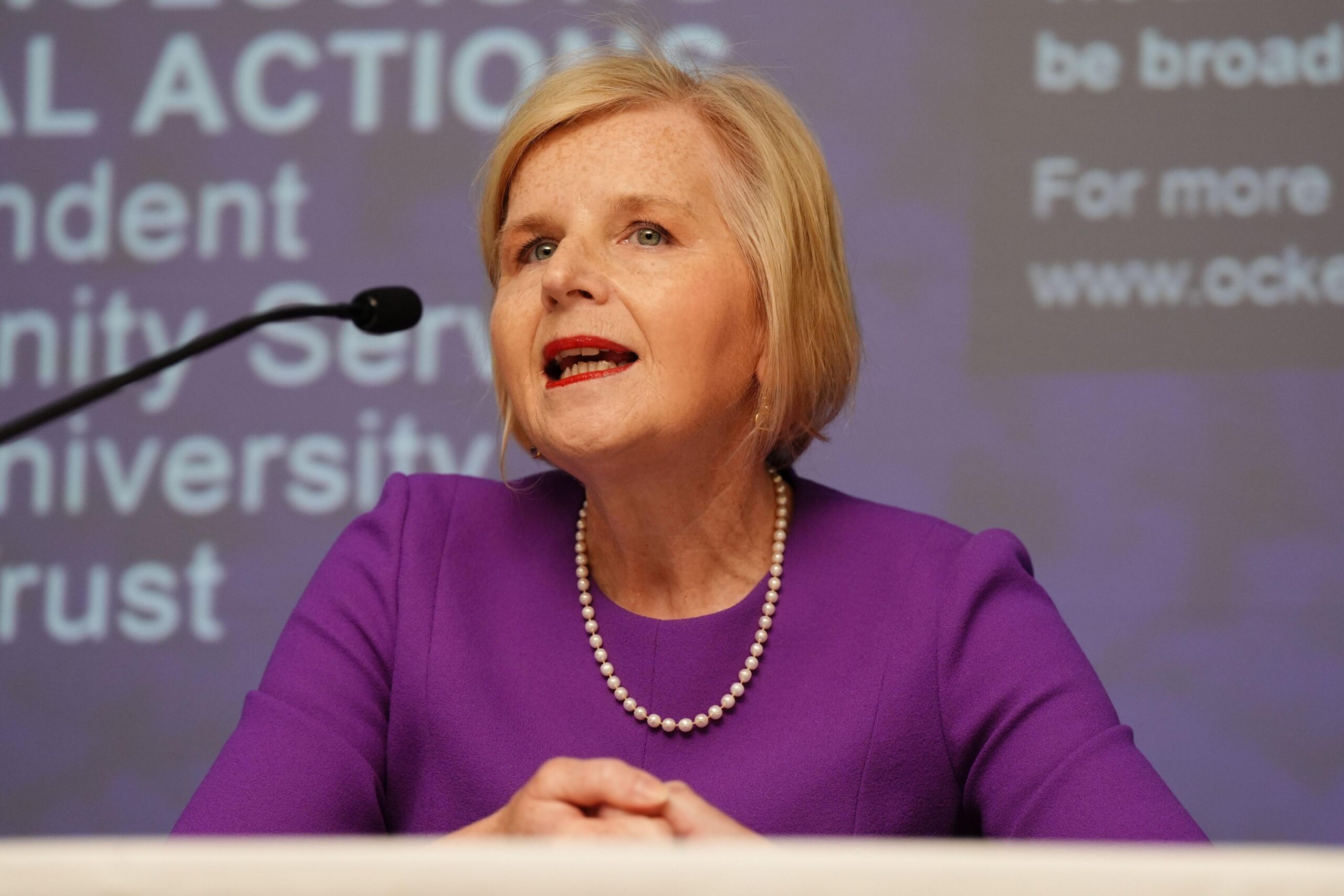 Donna Ockenden, who has short blonde hair, speaks into a microphone. Donna is wearing a purple dress and a white pearl necklace. A presentation can be seen on the screen behind her.