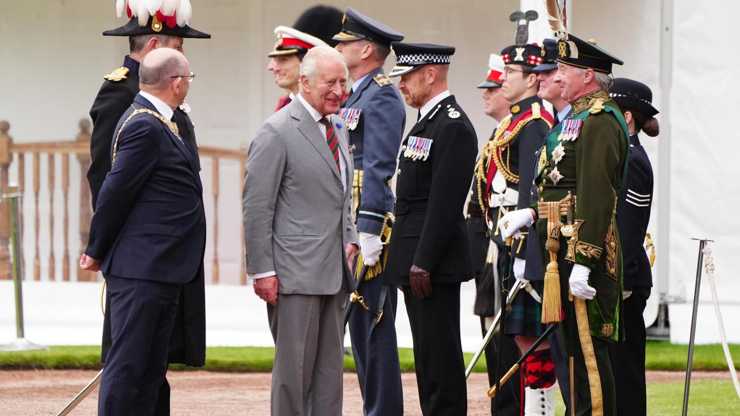 King Charles III wearing a grey suit and white shirt walks with the Lord Provost of Edinburgh Robert Aldridge, who wears glasses and a dark-coloured suit. The pair are greeted by uniformed figures.
