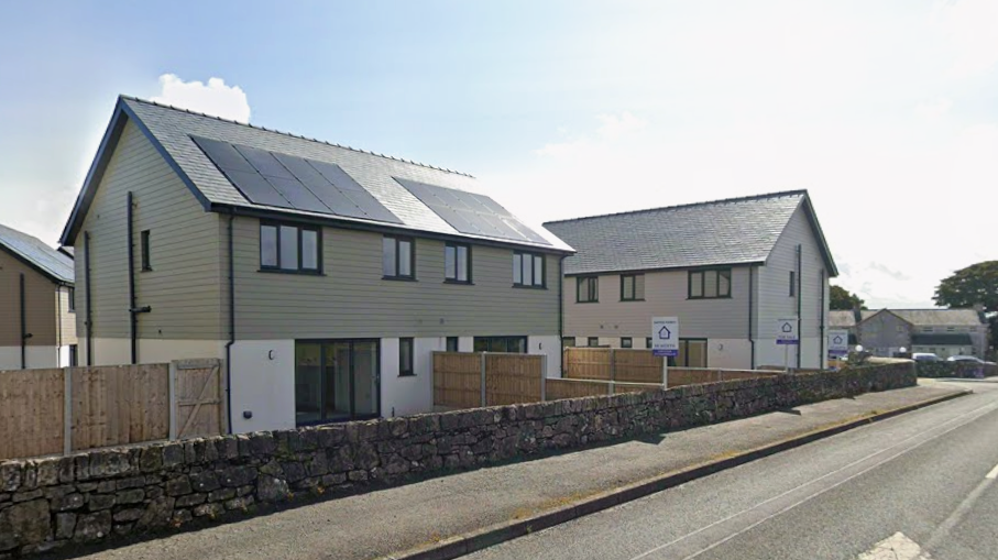 A street view of semi-detached homes lining a road. They have solar panels on the roof and green cladding, with wooden fences separating the homes. A number of for sale signs can be seen in the garden of the homes.