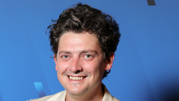A man with curly brown hair standing in front of a blue background wearing a cream suit jacket anfd smiling at the camera.