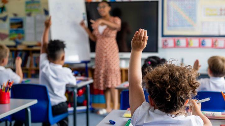 Children in a classroom sit at desks with hands raised while a teacher stands at the front beside a whiteboard, leading a lesson. Colourful posters decorate the walls and stationery items are visible on desks.