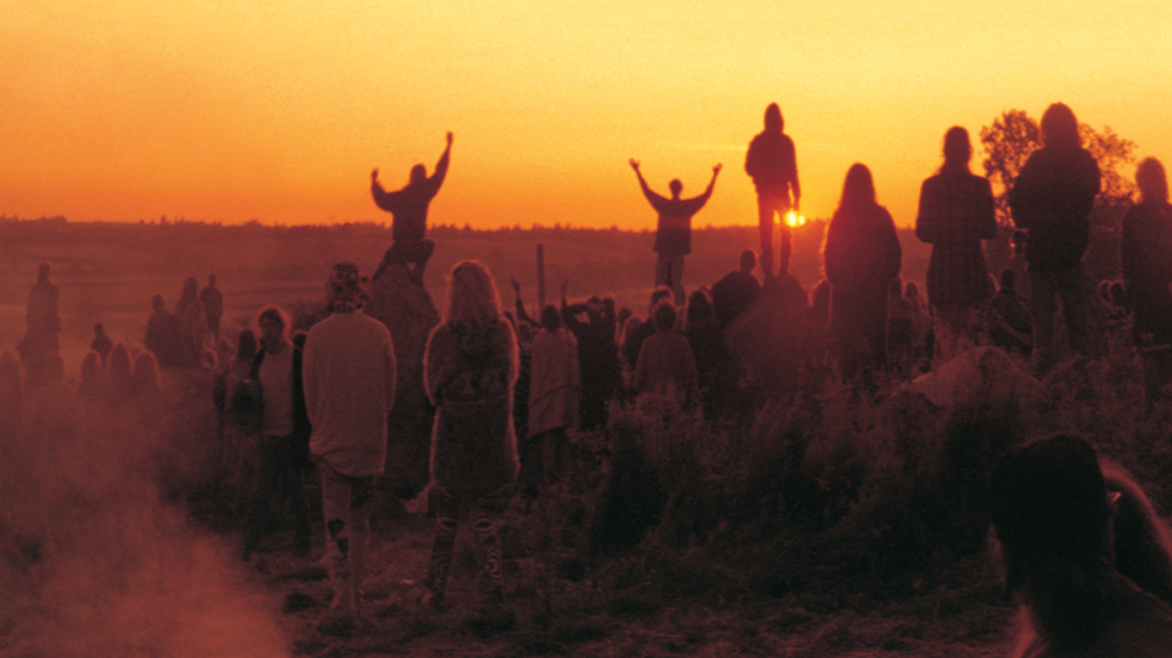 An image at sunrise with lots of people standing looking towards the horizon on a field in semi-darkness. There is an orange glow to the sky and two people are silhouetted in the distance with their arms in the air.