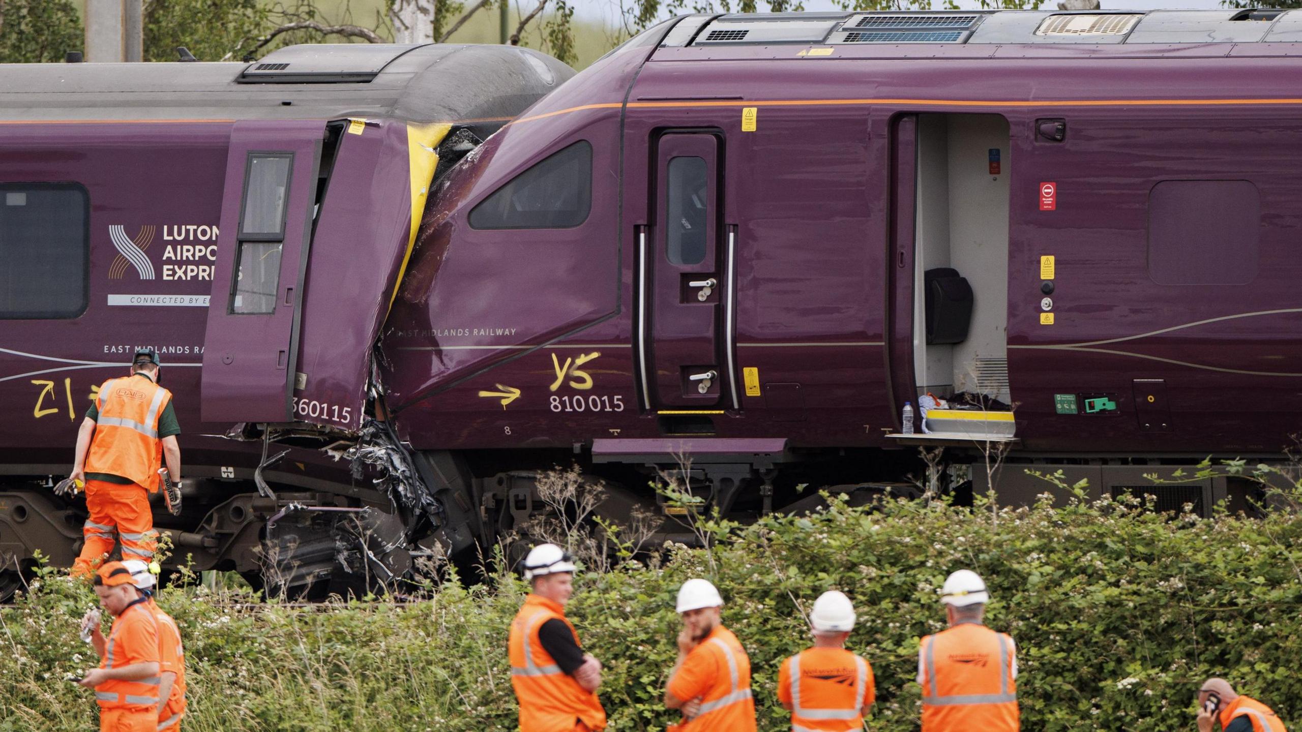 Two trains are seen crashed together with people wearing orange protective clothing standing alongside.