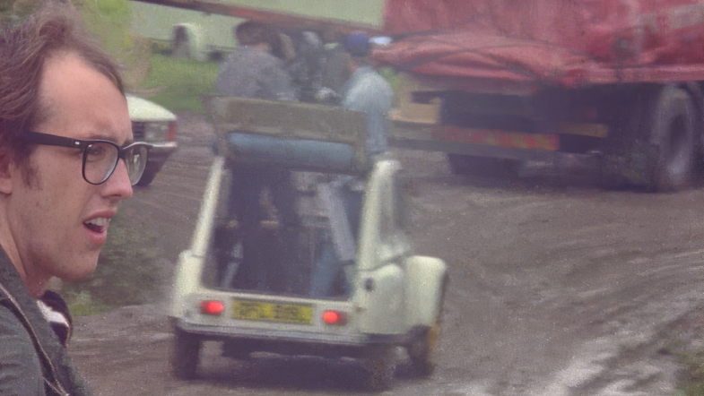 A vintage image of the camera crew in an open-topped car standing up through the roof with a large camera. Robin is in the foreground,  his face visible on the left. He is looking to the right, off camera. The ground is very muddy and there is a lorry with a red tarpaulin in the background.