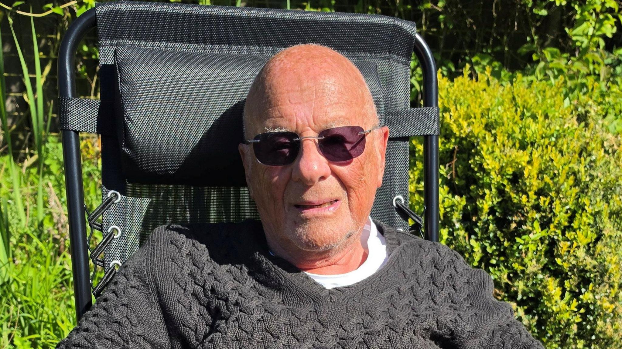 An older man wearing sunglasses and a grey jumper which is pulled over a white T-shirt. He is sitting in a chair outside with bushes behind him.