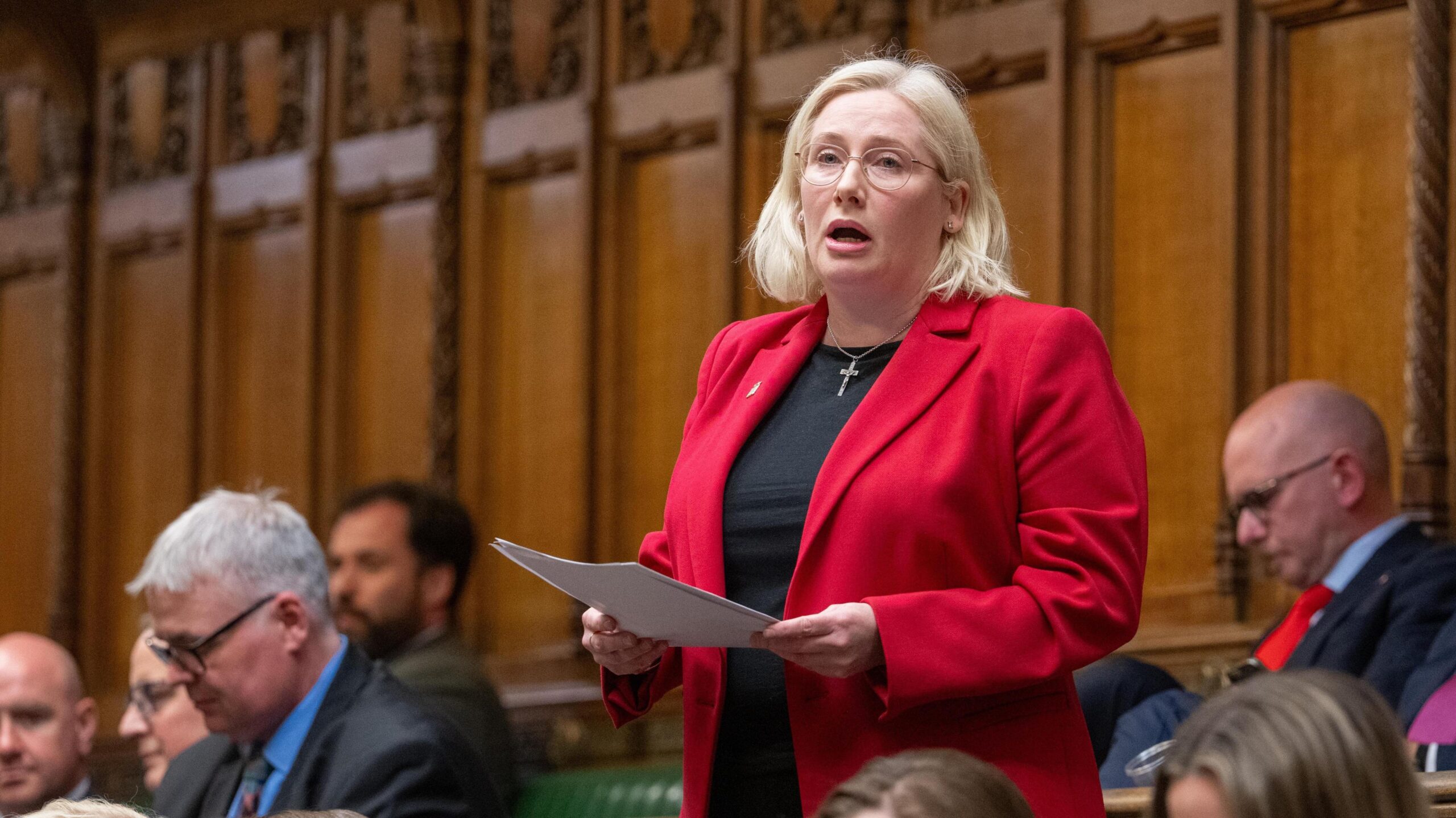 Emma Lewell MP stands giving a speech in front of the wood panelled walls of the House of Commons chamber surrounded by colleagues. She's wearing a crisp bright red blazer over a black top and has a silver crucifix necklace on. She's mid sentence holding papers in her hands in front of her.