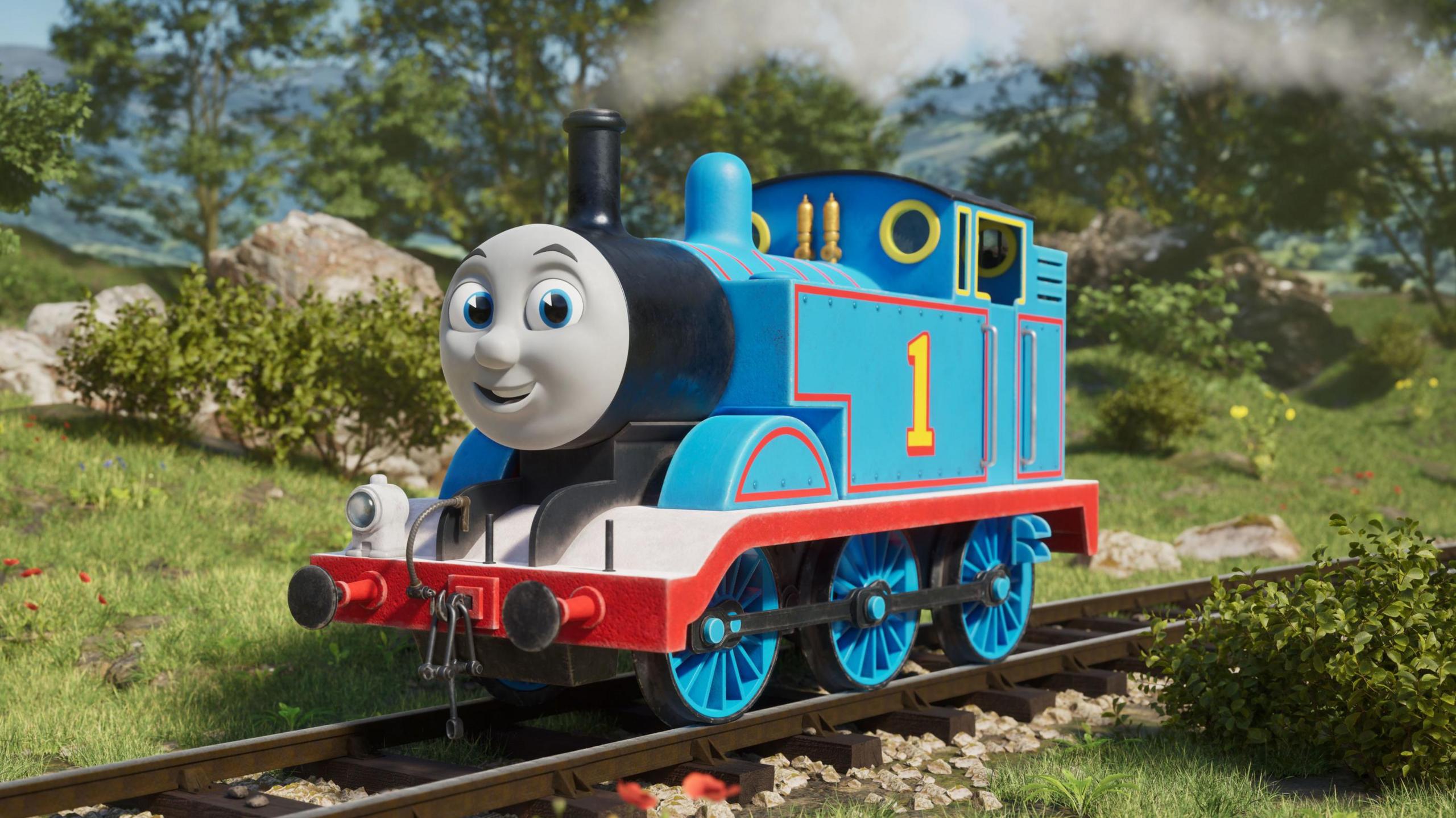 A blue train engine with a grey smiling face. It has a yellow number one on the side of it, and the track it is running along is surrounded by countryside.