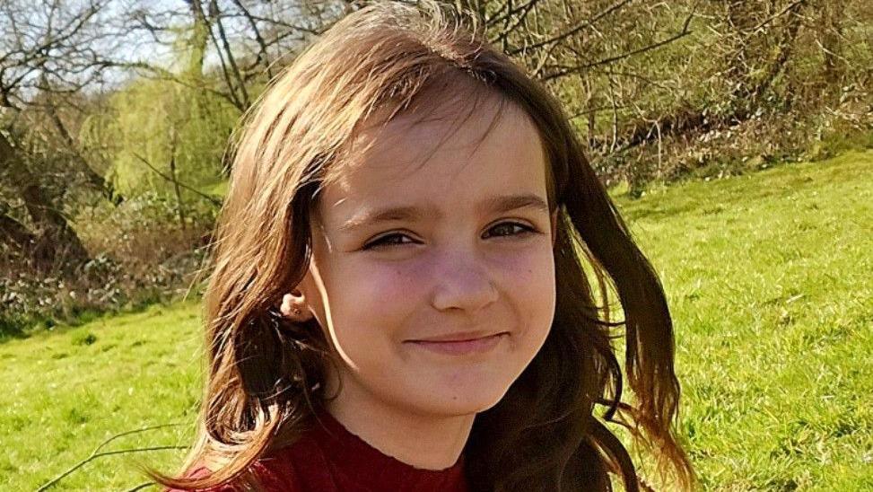 Aria Thorpe pictured outside. She has long dark hair and brown eyes. She is squinting in the sun and smiling. Grass and trees can be seen behind her.