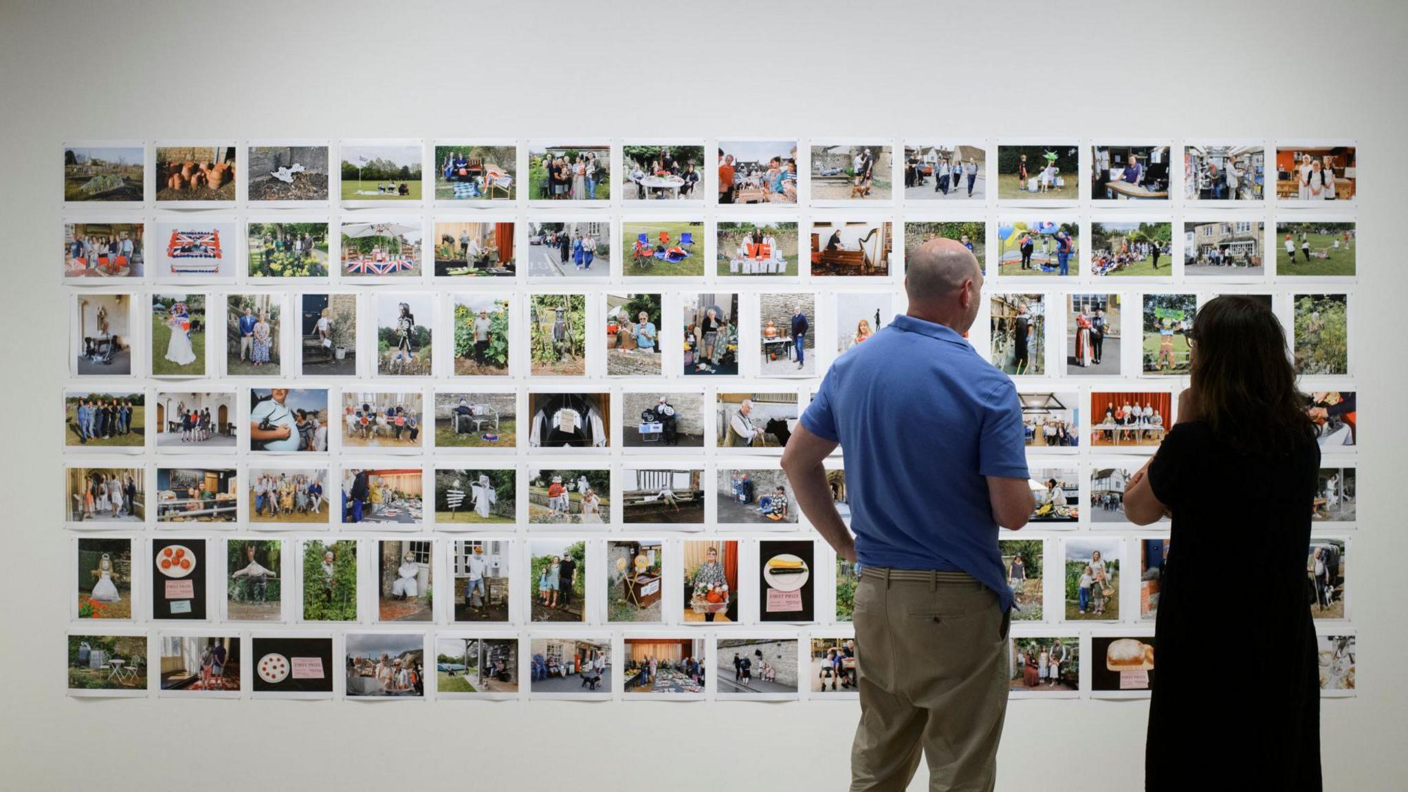 A man and a woman standing in front of a wall of photographs, with their backs to the camera.