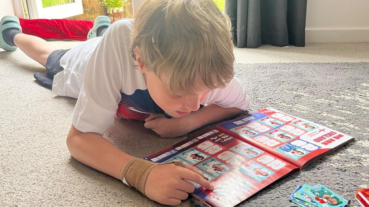 A young boy with blonde hair lies on the floor on his stomach, he is examining the England page of the sticker book and is pointing at a missing sticker. In front of him is a pile of stickers and a red tin with the World Cup logo on it, containing even more stickers.