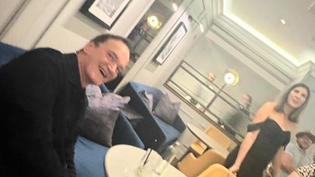 Quentin Tarantino sat on the left at a table. A woman in a black dress is walking through the venue.