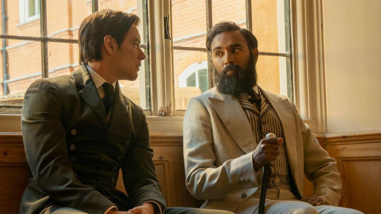 Louis Partridge and Himesh Patel in Enola Holmes sitting down talking to each other