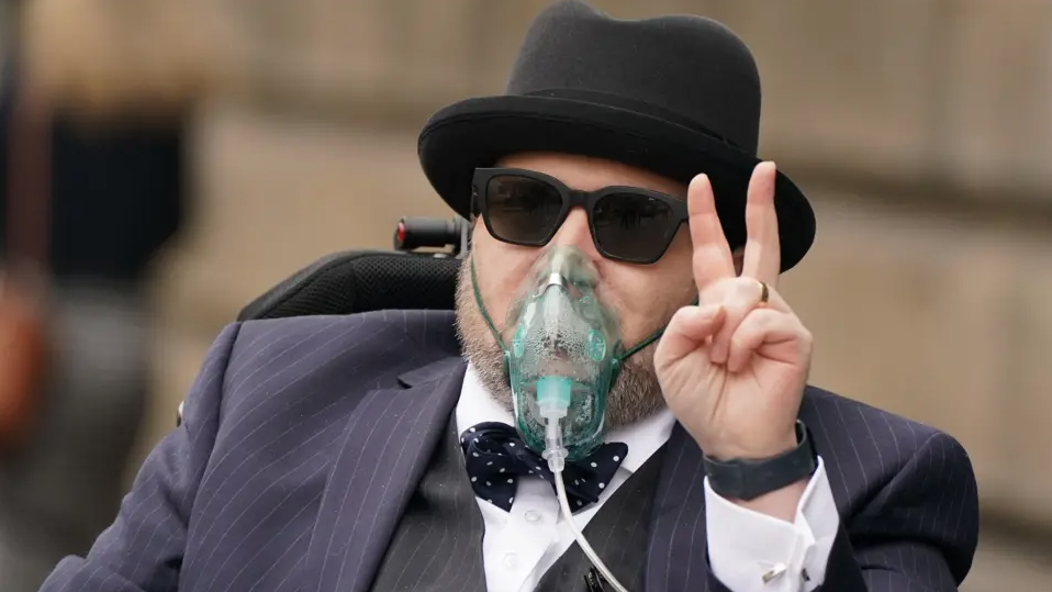 A man in a pin-striped suit and bow tie with trilby hat and sunglasses gives a victory salute
