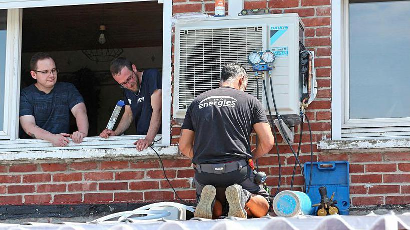 A man in black clothes with his back to the camera installs an air conditioning unit at a home in Mericourt, northern France on 19 June 19, 2026. From a window, two men look at him working