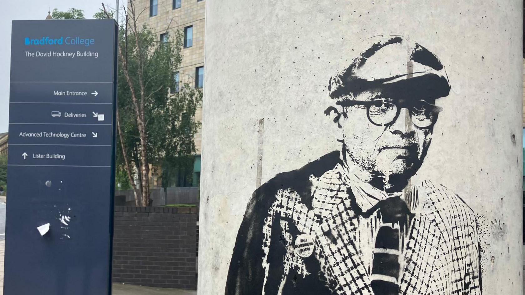 A graffiti image of David Hockney on a pillar with a sign for Bradford College behind