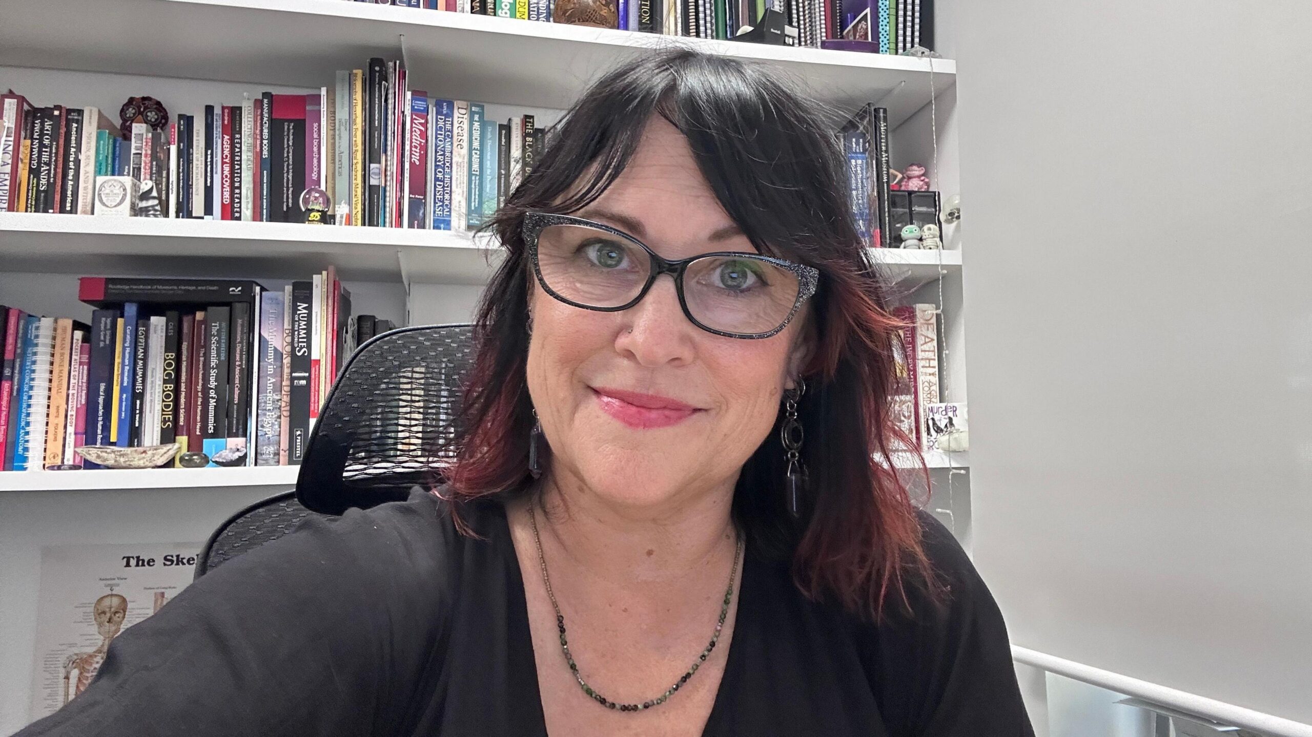 Dr Trish is sitting in front of a bookcase in a white office. She is wearing a black top, glasses, a necklace and has black hair with reddish/pinkish tips. She is wearing pink lipstick and smiling at the camera.