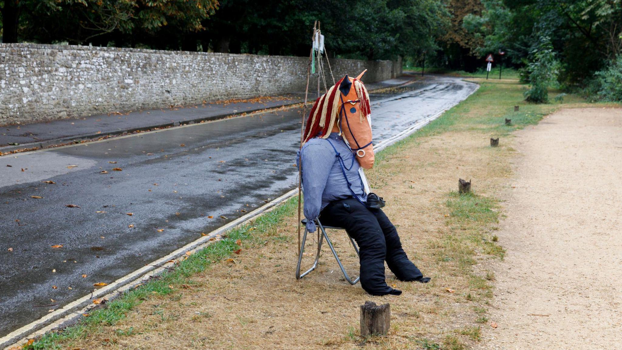 A scarecrow with a horse's head on a stool. The stool is on a grass verge at the side of a road.  A long stone wall and trees can be seen in the background.