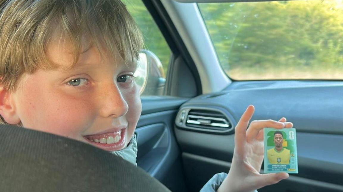 Jack, a young boy, sits in the front passenger seat of a car. He has a long blonde fringe and is smiling. In his hand is a 2026 Panini World Cup sticker showing Brazilian forward Vinicius Junior.