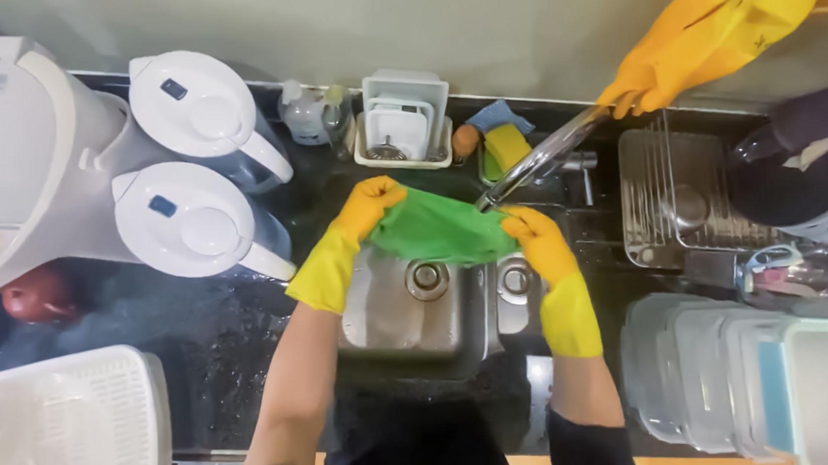 Point of view from a camera on a person's head showing them wearing yellow rubber gloves and holding a green washcloth under the tap in a kitchen sink