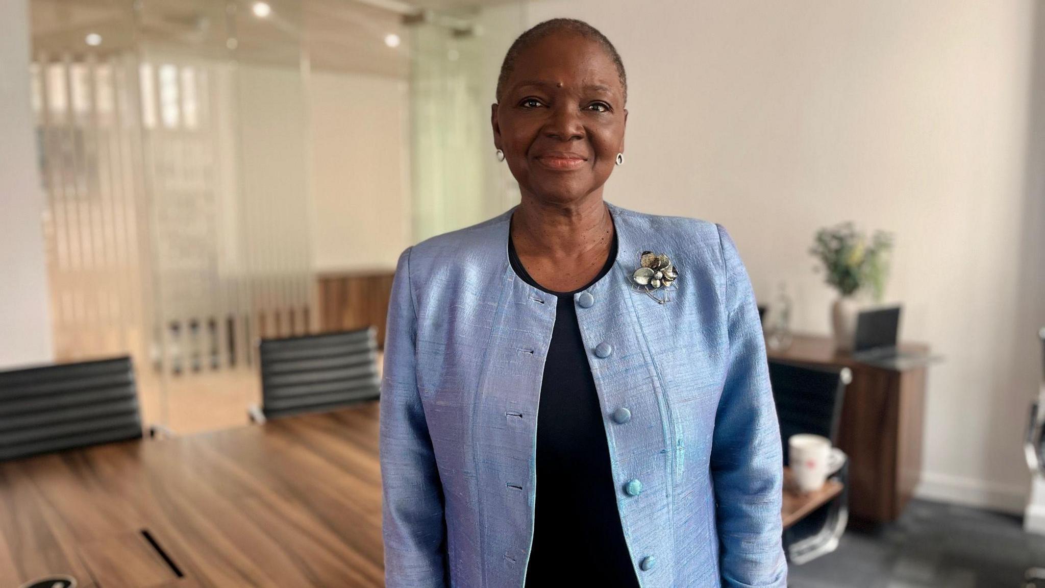 A photo of Baroness Amos from the waist up. She wears a baby blue blazer with a broach, has cropped hair and is wearing silver earrings. The background behind her is slightly blurred, but a large table and chairs can be seen.
