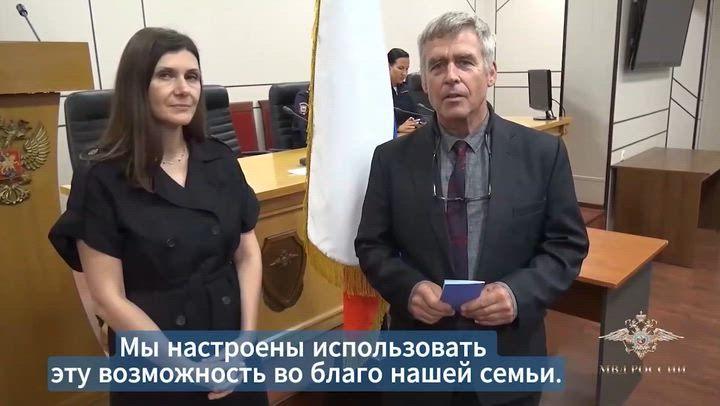 Two people in formal clothing stand at the front of a courtroom, one holding a small blue booklet and folded glasses. A light-coloured flag on a pole stands between them. Wooden desks, chairs, and a lectern with an emblem are visible behind. A person sits at a desk in the background. A crest appears on the right side of the image.